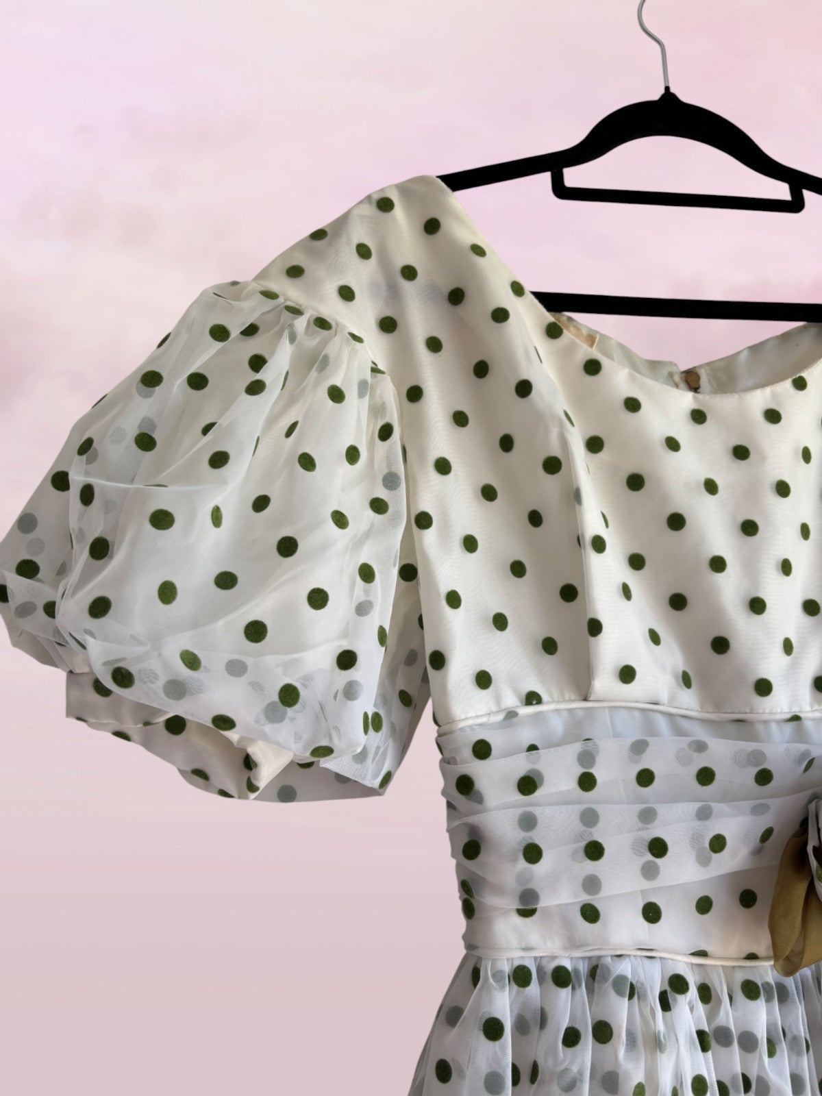 White blouse with green polka dots on a hanger against a pink background