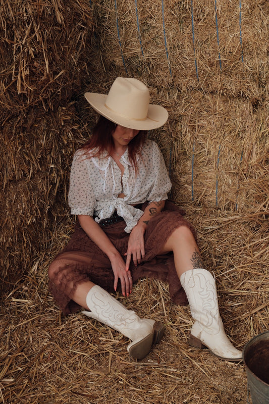 Person wearing a cowboy hat, white shirt, brown skirt, and white boots sitting on hay.
