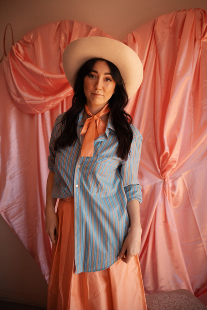 Woman wearing a blue striped shirt and pink skirt with a beige hat in front of pink curtains.