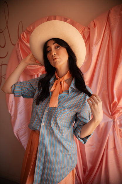 Woman wearing a blue striped shirt, orange scarf, and wide-brimmed hat against a pink curtain.