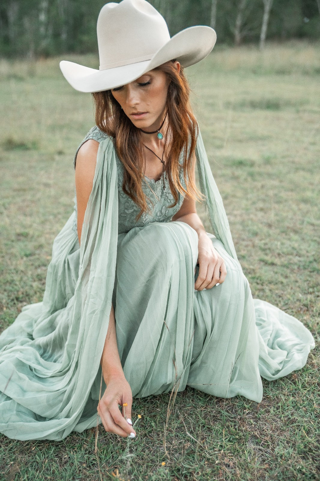 Woman in a long light green dress and cowboy hat sitting on the grass.