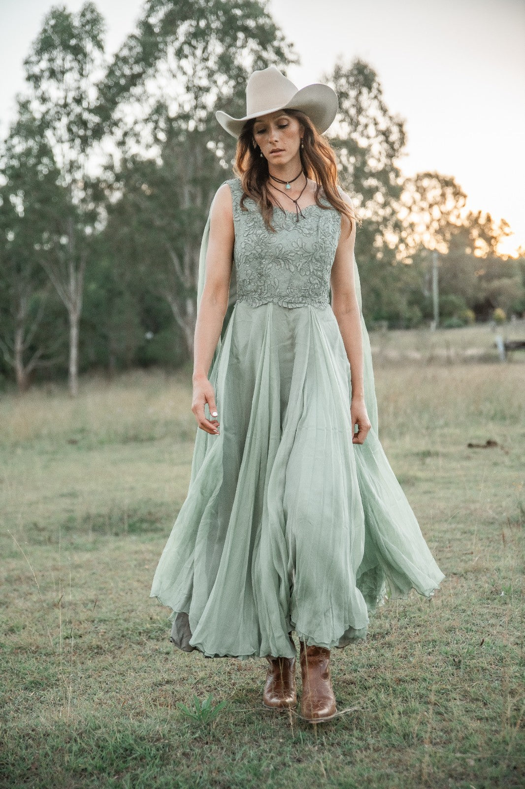 Woman in a long light green dress and cowboy hat standing in a field.