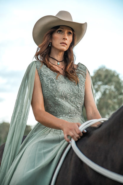 Woman in a light green dress and cowboy hat riding a horse outdoors.