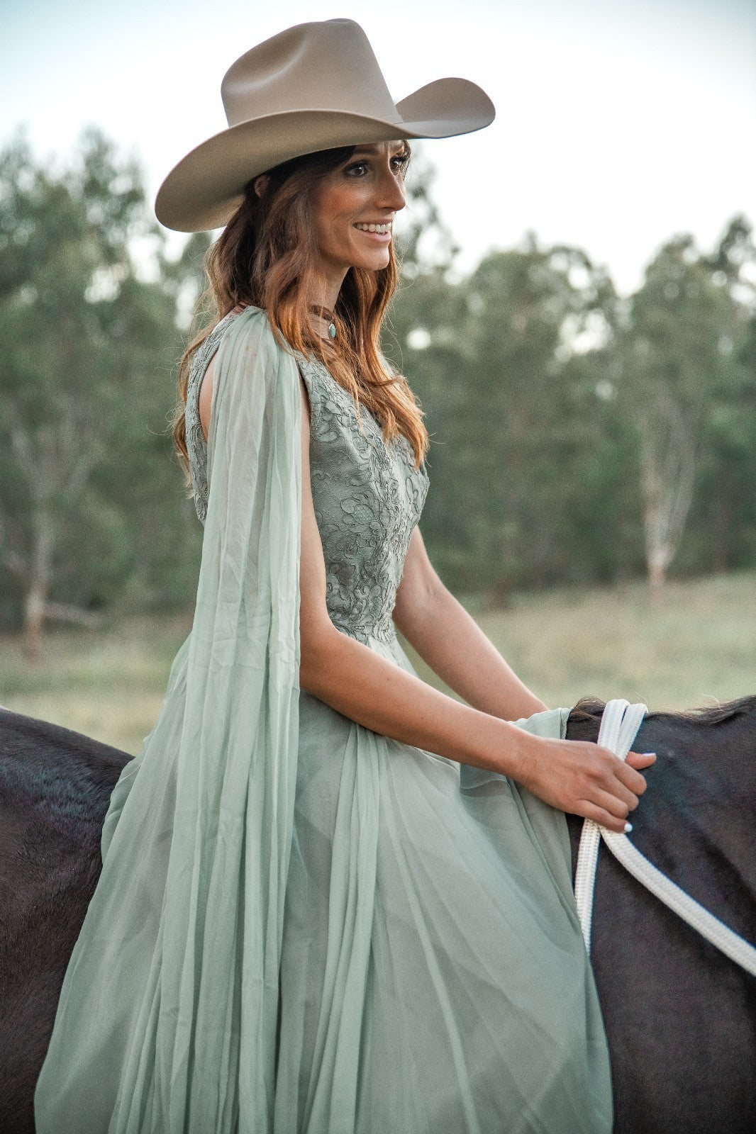 Woman in a light green dress and beige cowboy hat sitting on a horse with a blurred natural background