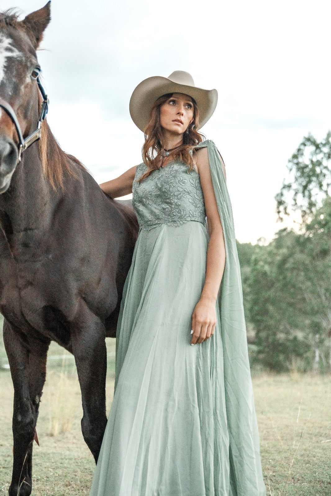 Woman in a light green dress and cowboy hat standing next to a horse in an outdoor setting.