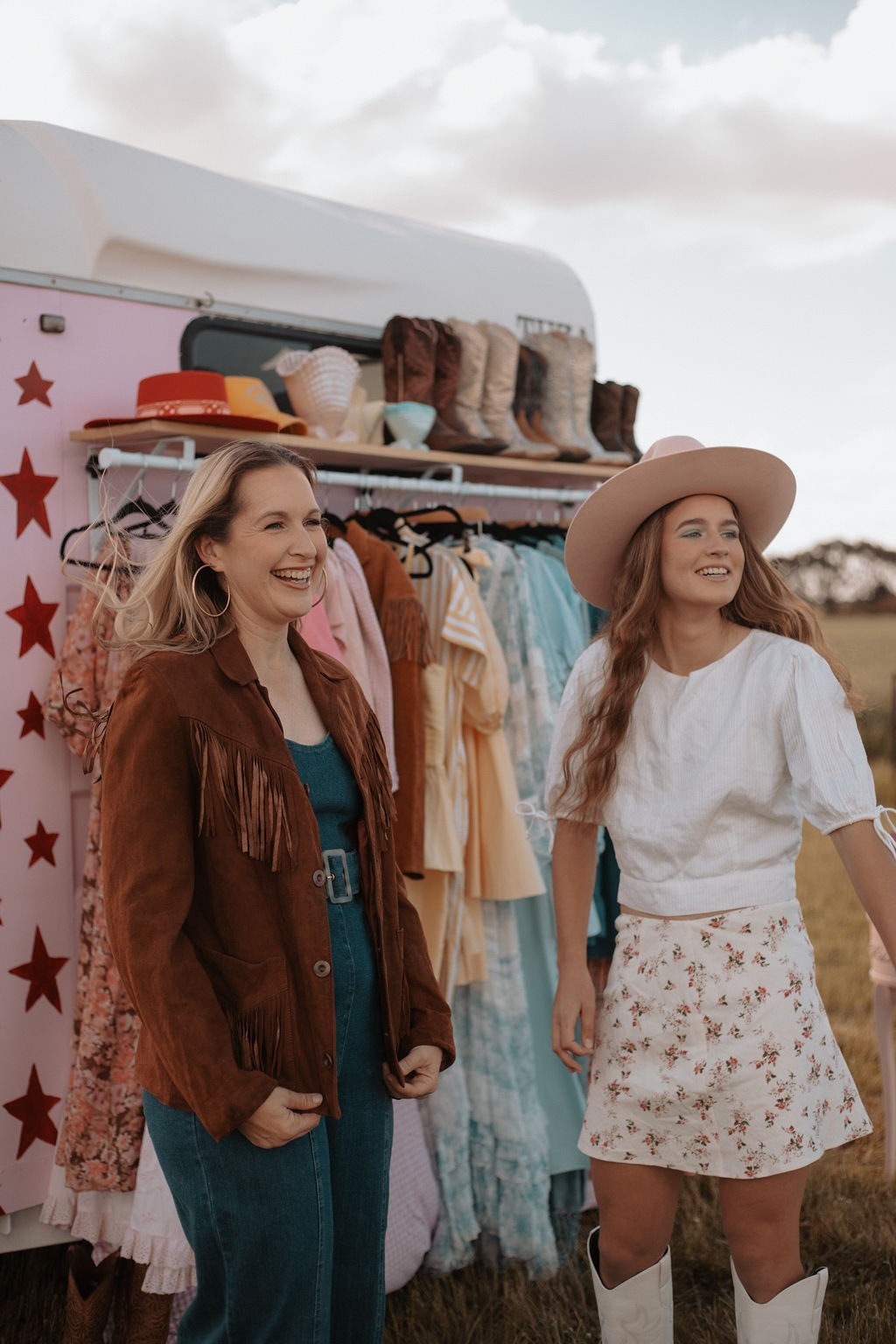 Two women standing in front of a vintage-style trailer with clothing racks, smiling.