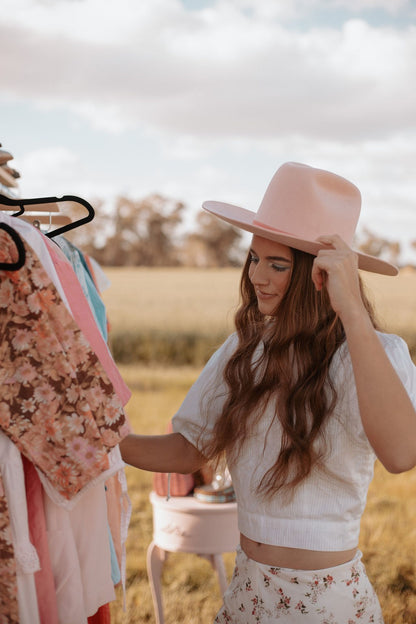 Woman in a field wearing a pink hat and white outfit, standing next to a rack of clothes.