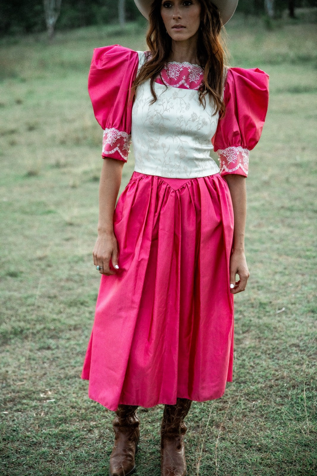 Woman wearing a pink dress with white top and floral embroidery, standing in a grassy field.