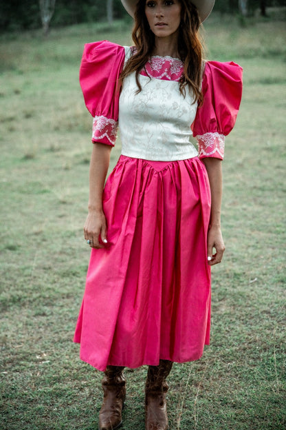 Woman wearing a pink dress with white top and floral embroidery, standing in a grassy field.