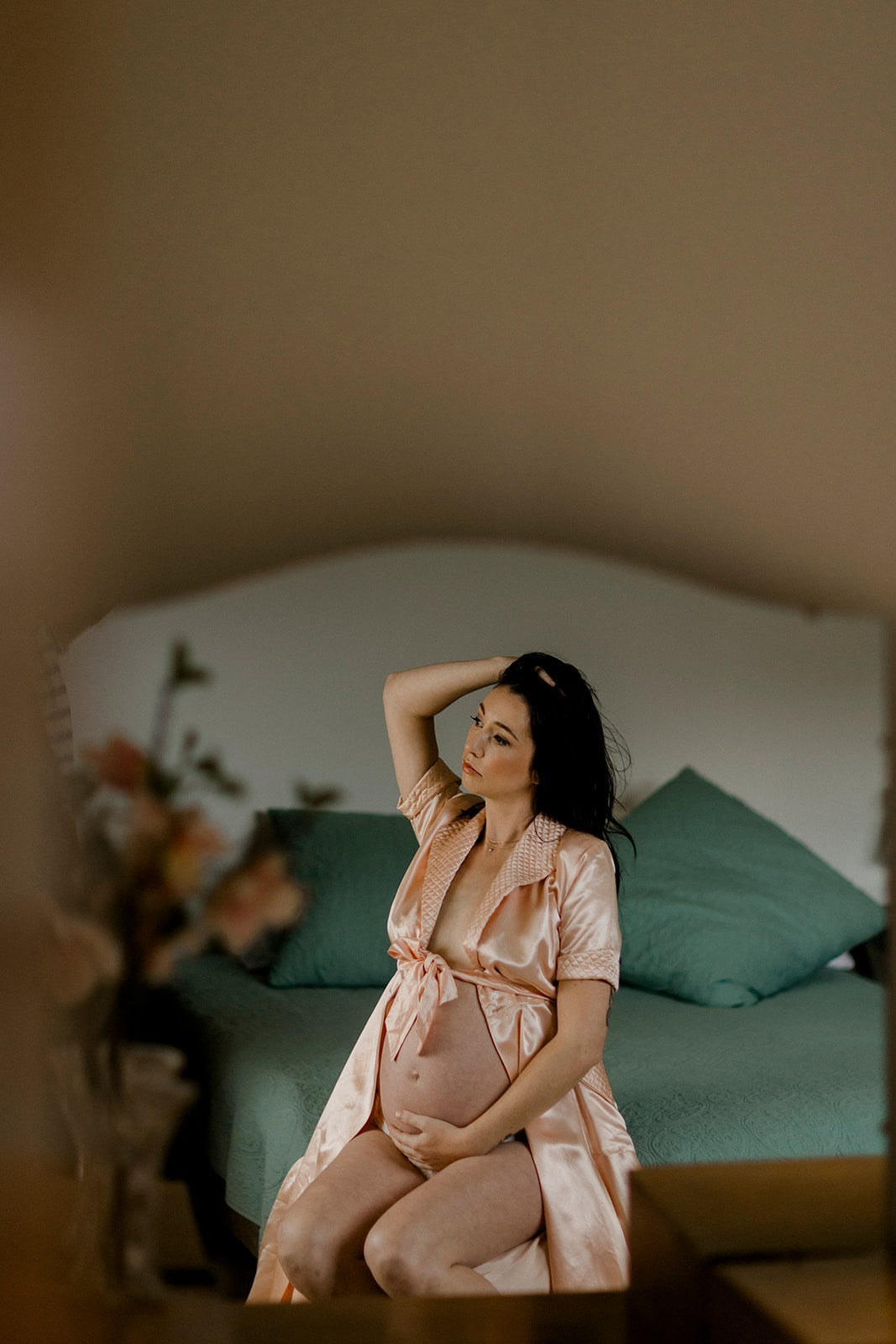 Woman in a pink robe sitting on a bed with teal pillows, surrounded by flowers.
