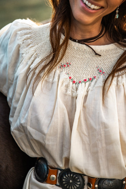 Woman wearing a white blouse with floral embroidery and a belt, standing outdoors.