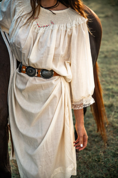 Person wearing a white dress with lace details and a belt, standing outdoors.