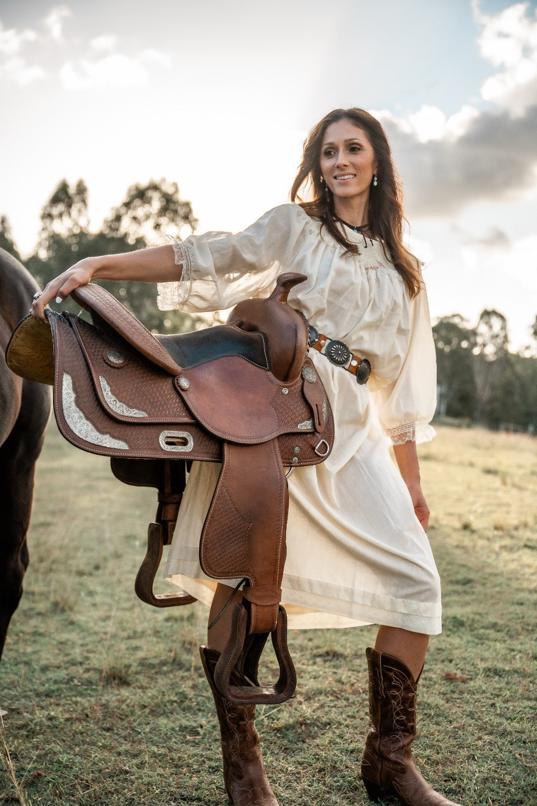 Woman in a white dress and brown boots holding a horse saddle in an outdoor setting.