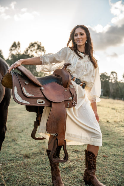 Woman in a white dress and brown boots holding a horse saddle in an outdoor setting.