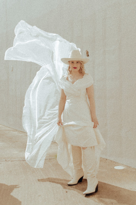 Woman in a white dress and hat holding a flowing white fabric against a neutral background