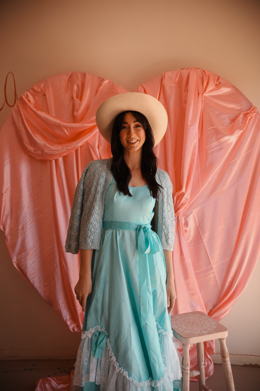 Woman in a light blue dress and beige hat standing in front of a pink curtain