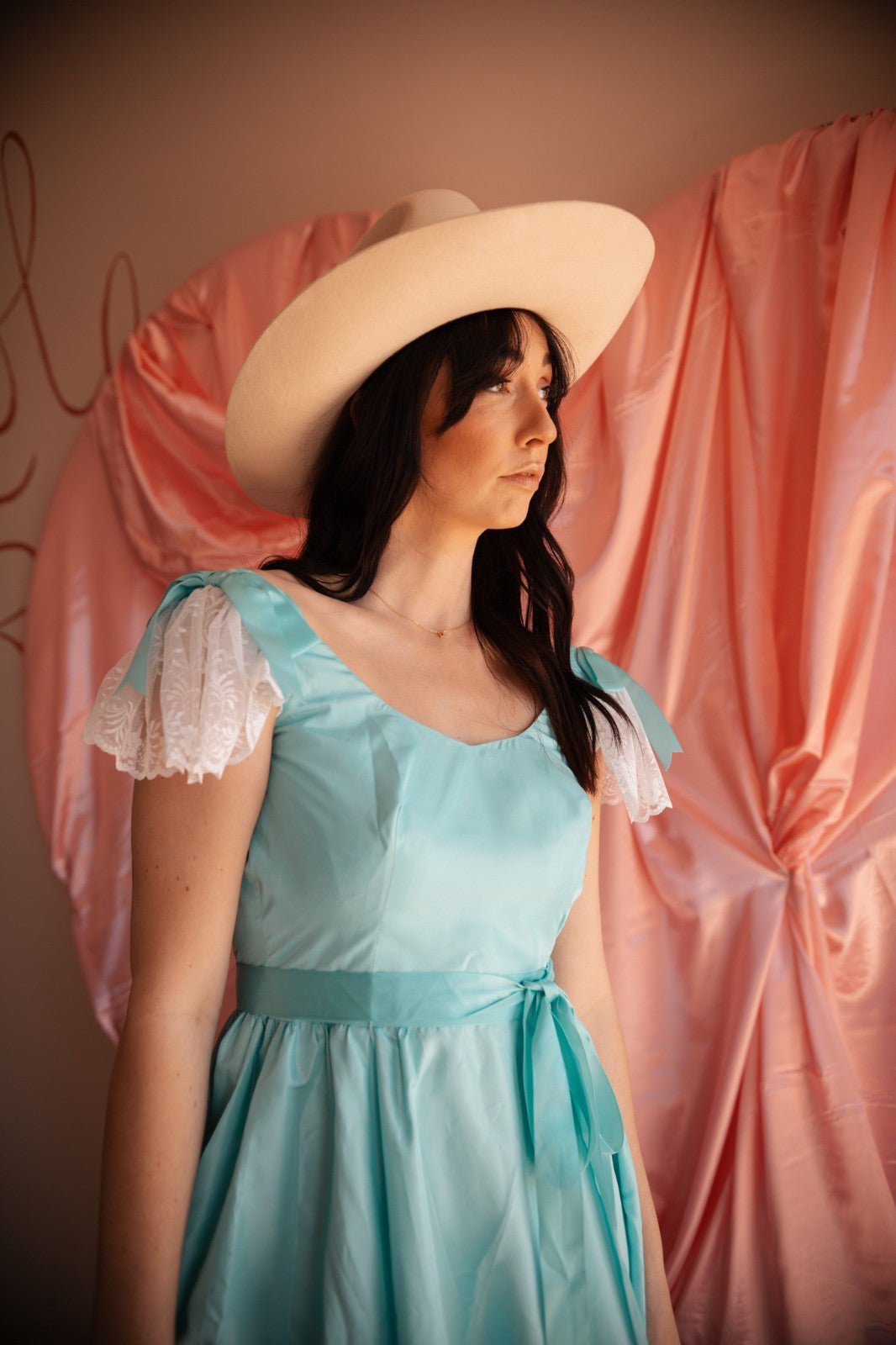 Woman wearing a light blue dress and beige hat against a pink curtain backdrop