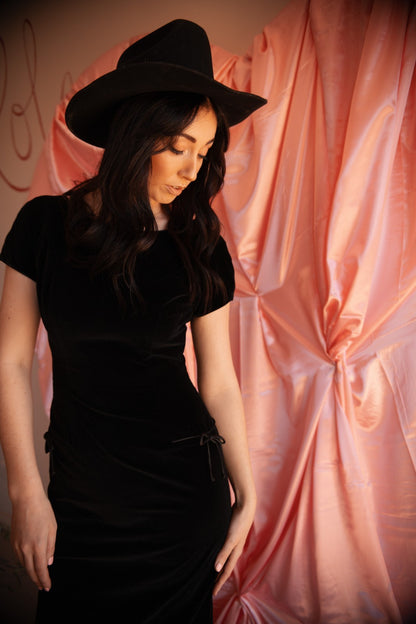 Woman wearing a black dress and hat against a pink draped fabric background