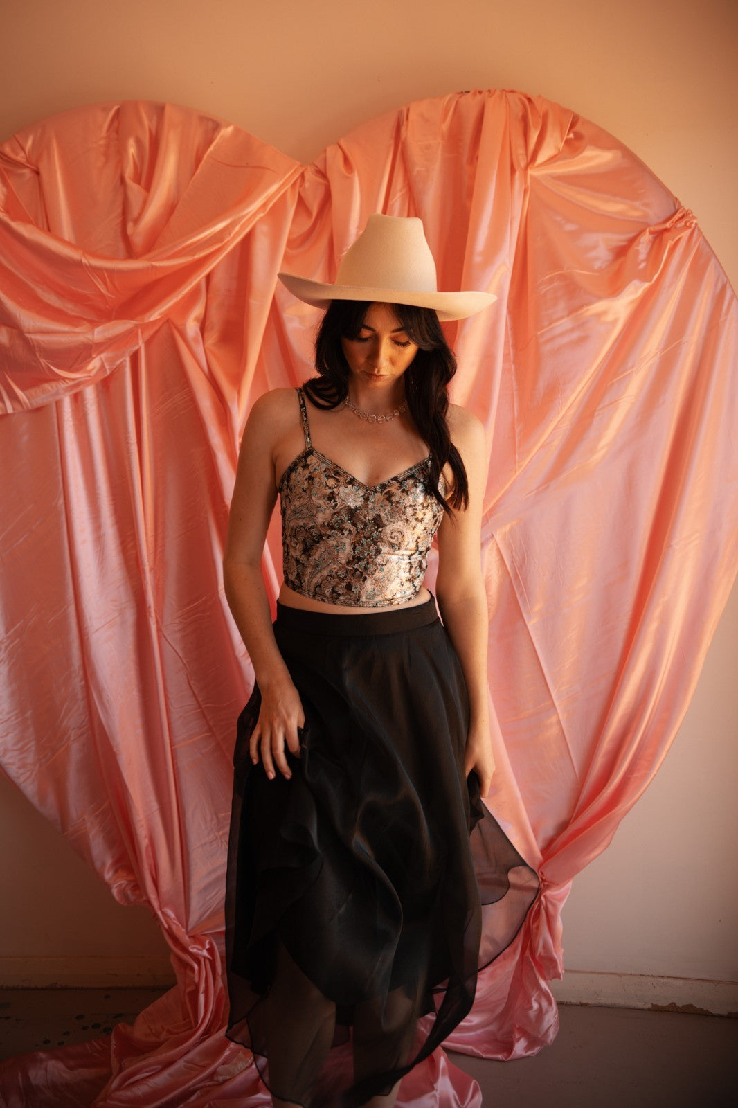 Woman wearing a floral top and black skirt with a beige cowboy hat in front of pink draped fabric.