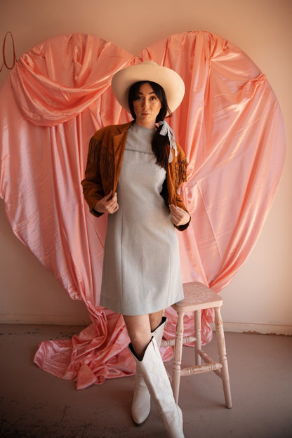 Woman in a light blue dress and white boots standing on a stool against a pink heart-shaped curtain.
