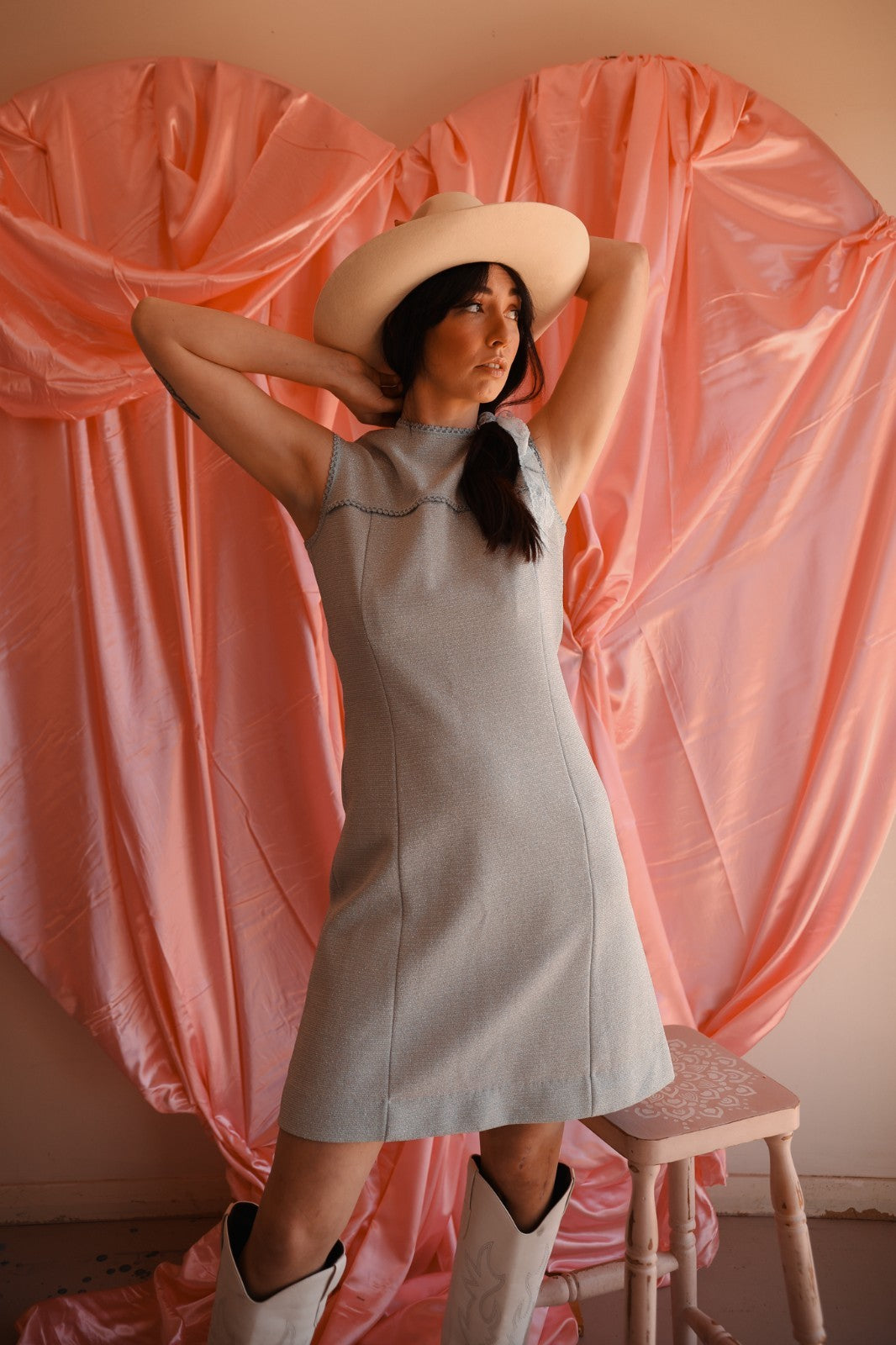 Woman in a light blue dress and white hat posing against a pink curtain backdrop.