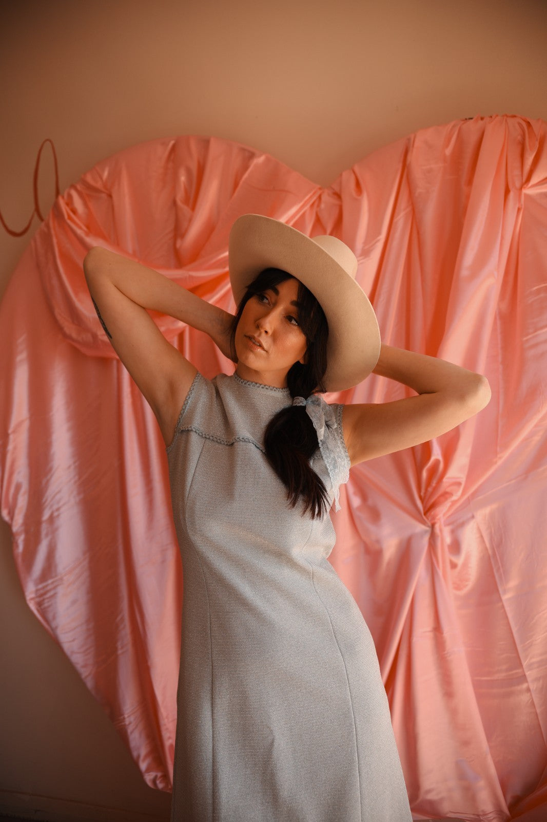 Woman wearing a light blue dress and beige hat against a pink heart-shaped curtain.