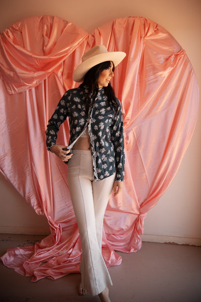 Woman in a floral shirt and white pants standing in front of a large pink heart-shaped curtain.