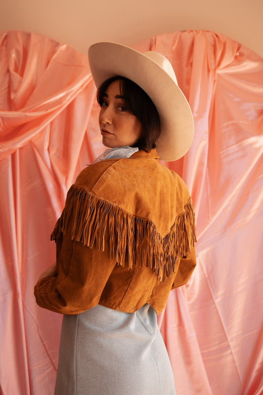 Person wearing a fringed jacket and wide-brimmed hat against a pink curtain backdrop
