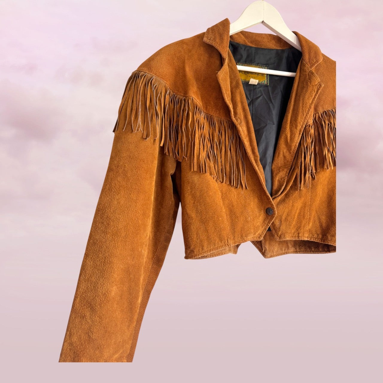Brown suede jacket with fringes on a hanger against a light purple background