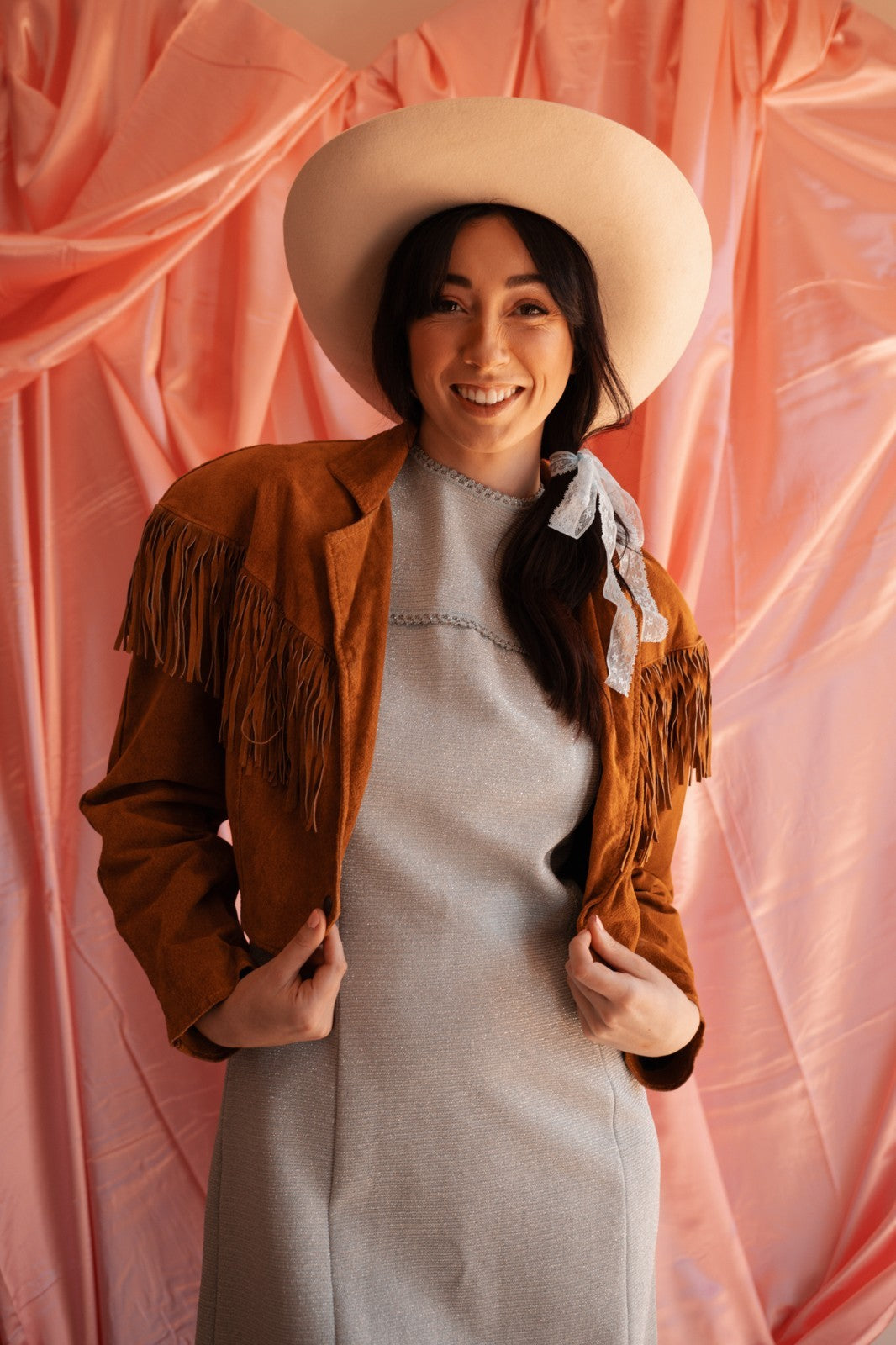 Woman wearing a brown fringed jacket and white hat against a pink background