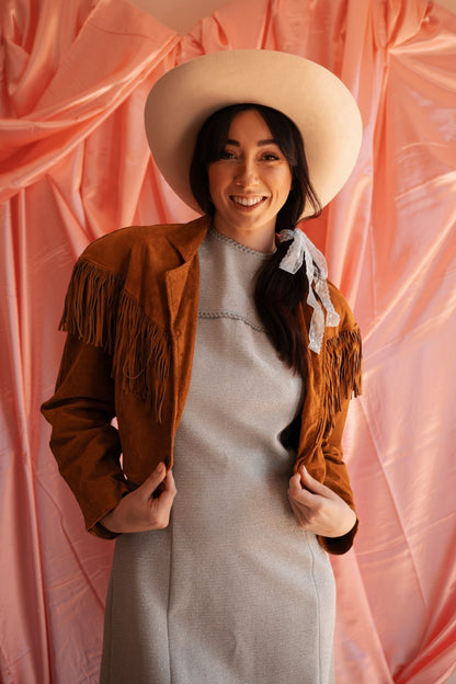 Woman wearing a brown fringed jacket and white hat against a pink background