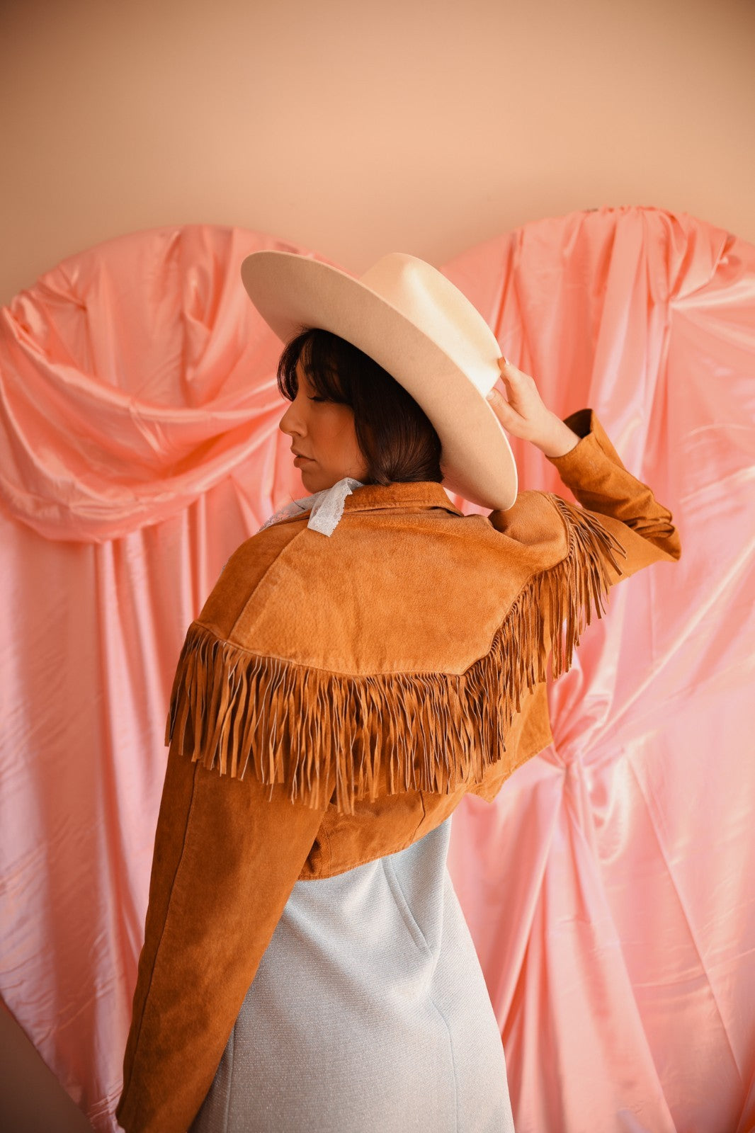 Person wearing a brown fringed jacket and white hat against a pink curtain background