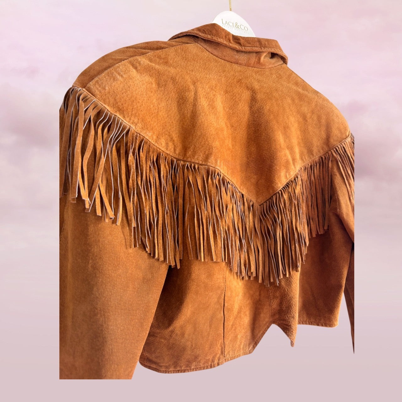 Brown suede jacket with fringes on a hanger against a light pink background