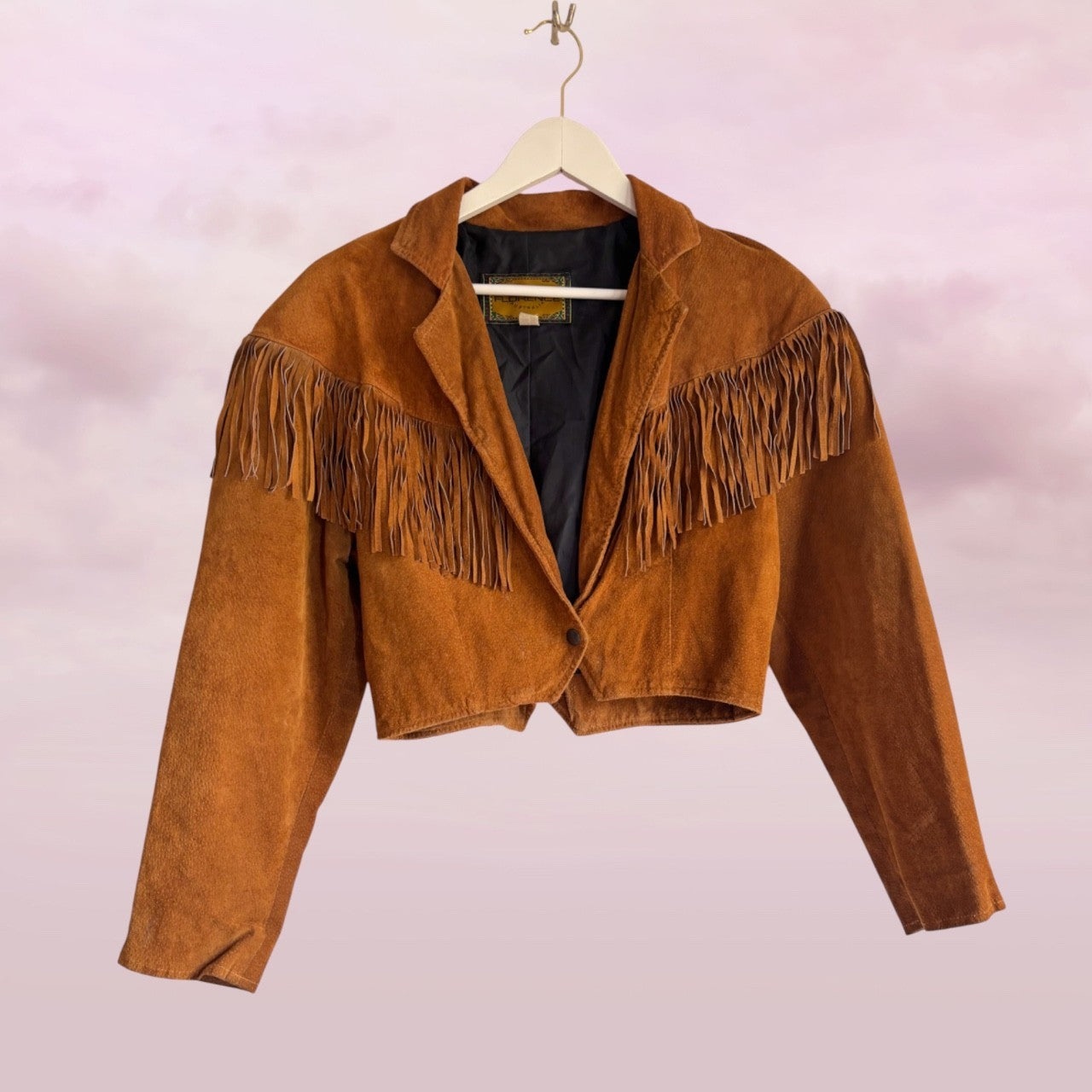 Brown suede jacket with fringe on a hanger against a light pink background