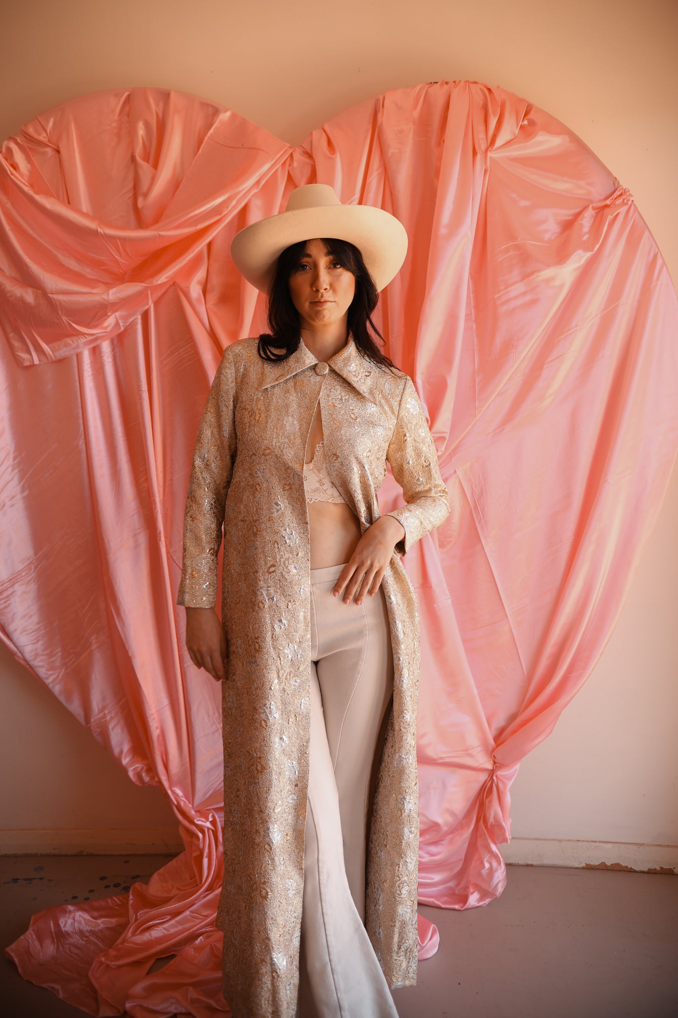Person in a beige outfit with a hat standing in front of pink draped fabric