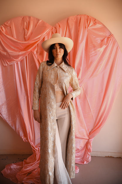 Person in a beige outfit with a hat standing in front of pink draped fabric