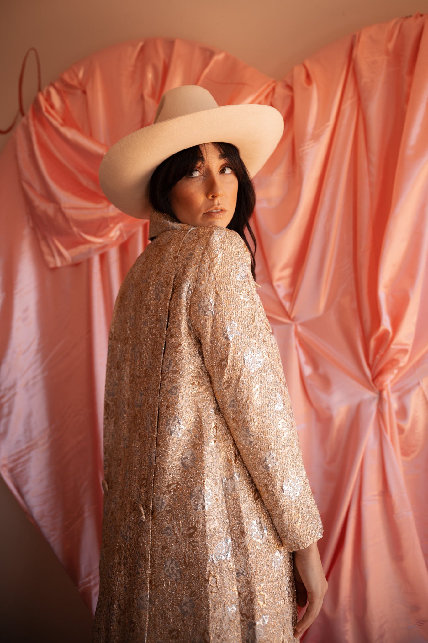 Person wearing a beige hat and patterned coat in front of pink draped fabric