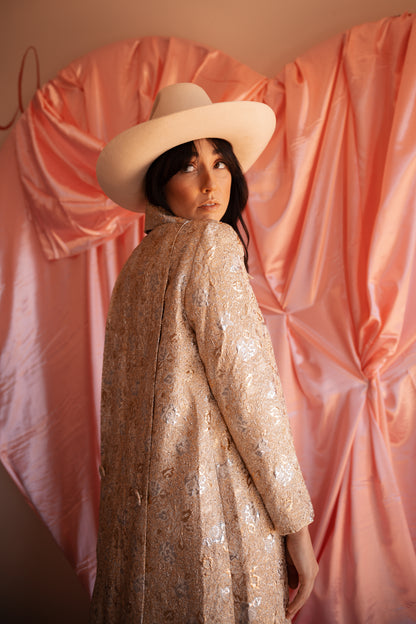 Person wearing a beige hat and patterned coat in front of pink draped fabric