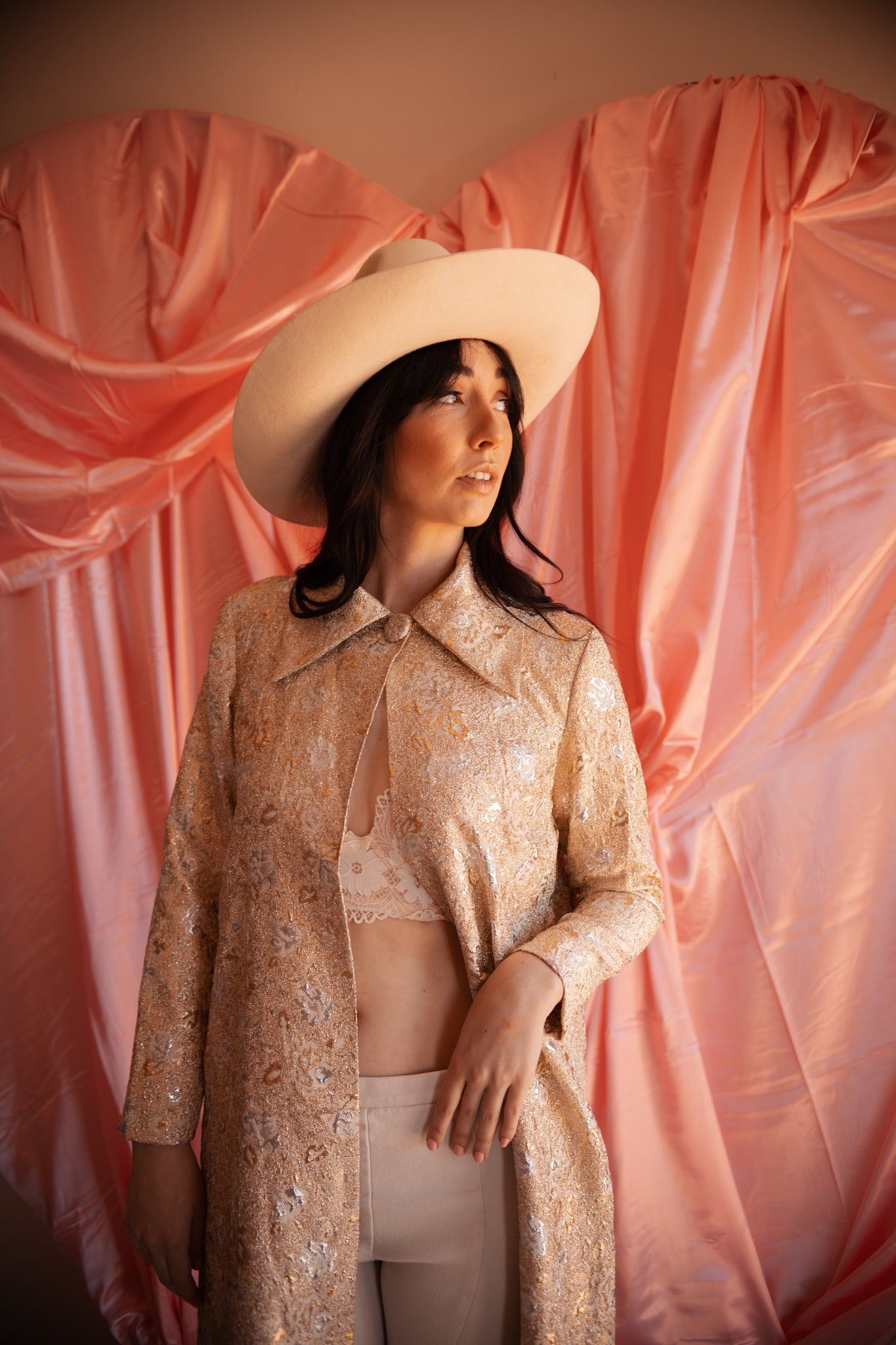 Woman wearing a patterned coat and wide-brimmed hat against a pink draped backdrop