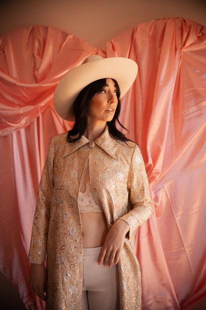 Woman wearing a patterned coat and wide-brimmed hat against a pink draped backdrop
