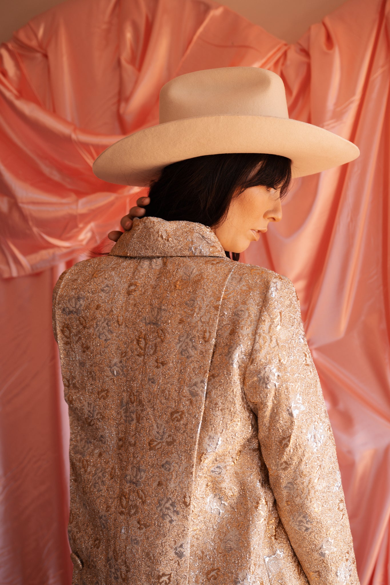 Person wearing a patterned coat and wide-brimmed hat against a pink fabric background