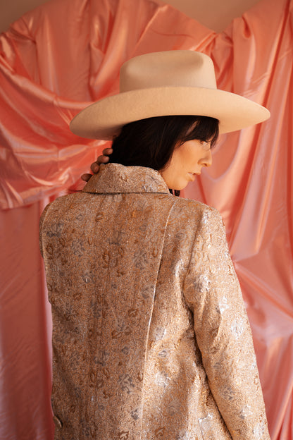 Person wearing a patterned coat and wide-brimmed hat against a pink fabric background