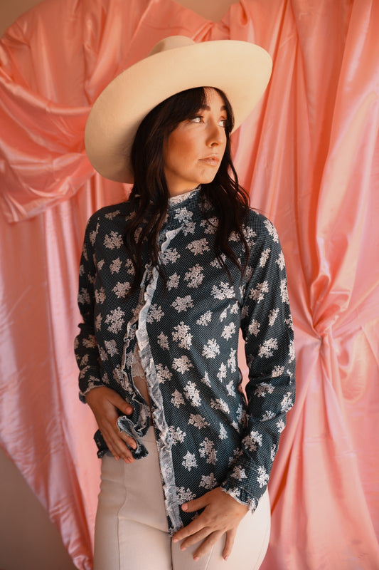 Woman wearing a floral blouse and wide-brimmed hat against a pink curtain backdrop