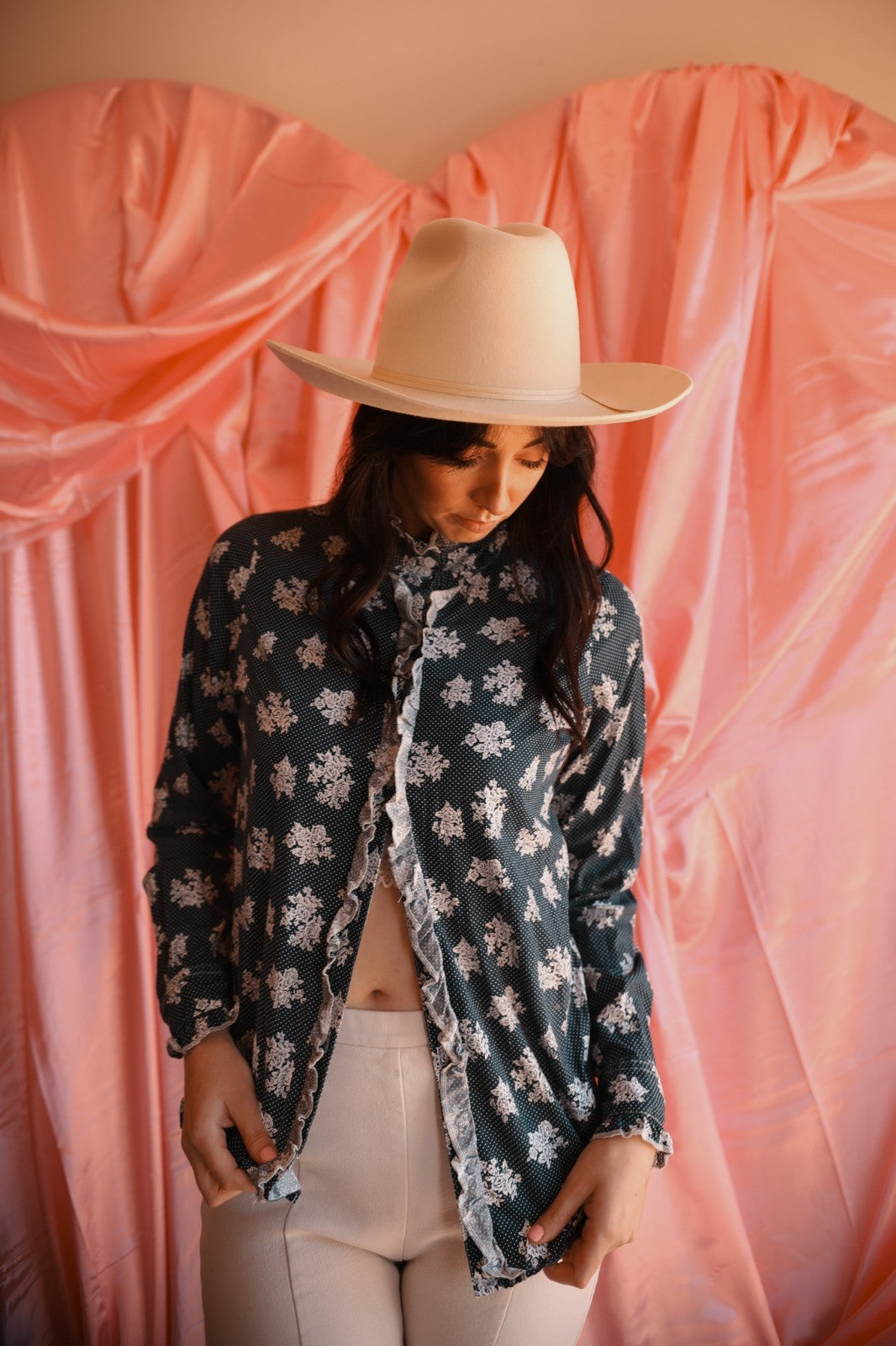 Person wearing a floral blouse and beige hat against a pink curtain backdrop