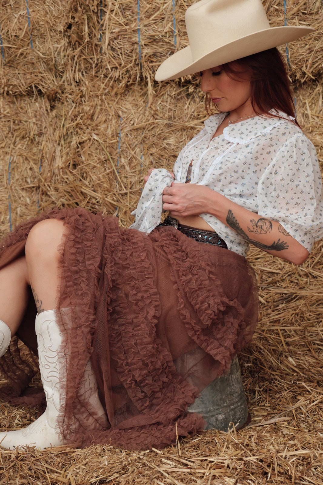 Woman in cowboy hat and boots sitting on hay with a brown skirt