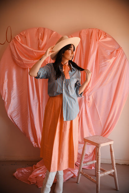 Woman in a peach dress and denim shirt standing in front of a pink curtain backdrop.