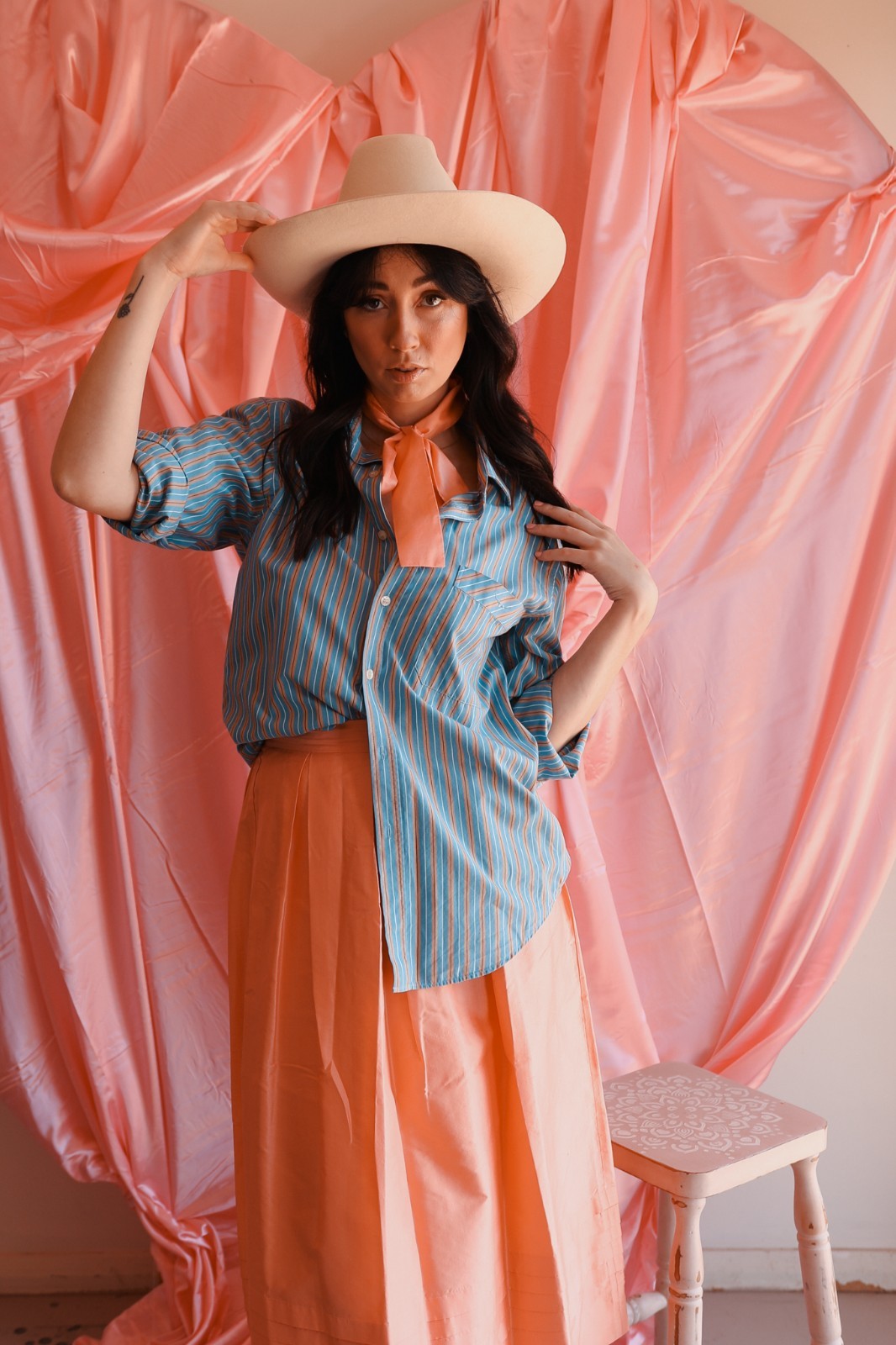 Woman wearing a blue striped shirt and orange skirt with a beige hat against a pink curtain backdrop.