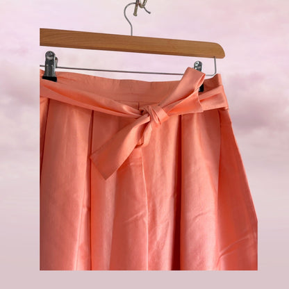 Coral pink skirt with a belt on a wooden hanger against a light pink background