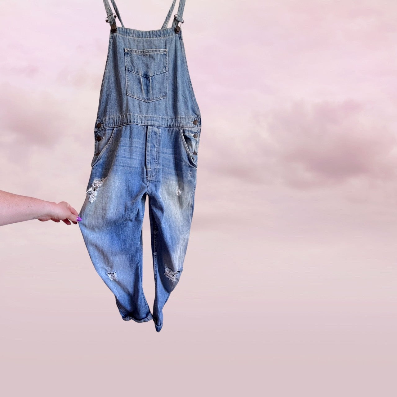 Zara preloved distressed Denim overalls held by a hand against a pink background front view 