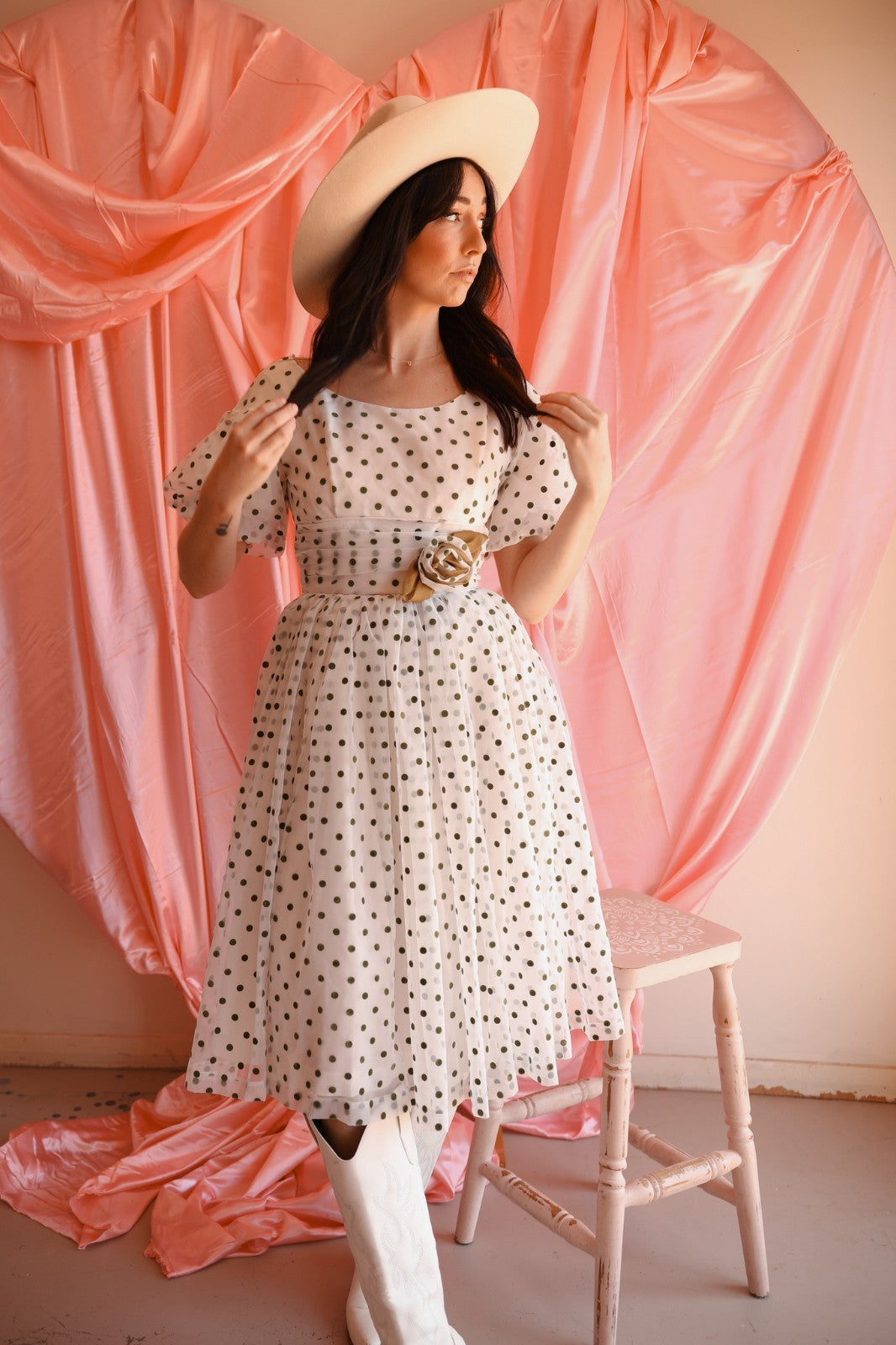 Woman in a polka dot dress and wide-brimmed hat standing in front of pink draped fabric.
